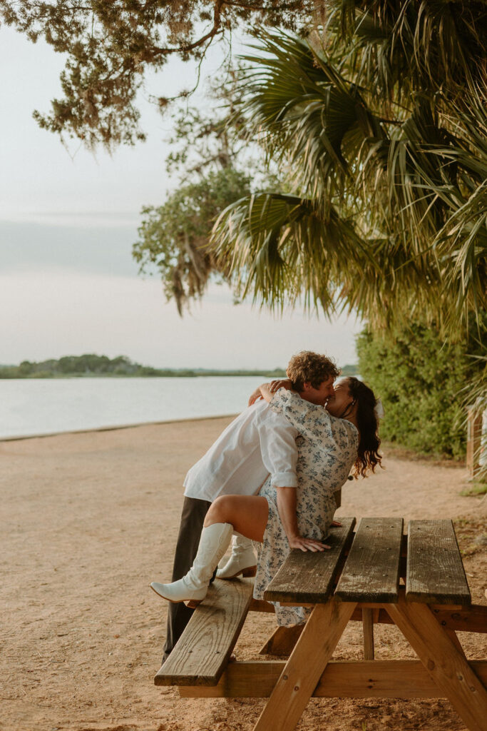 man leaning over woman kissing on a bench at the beach