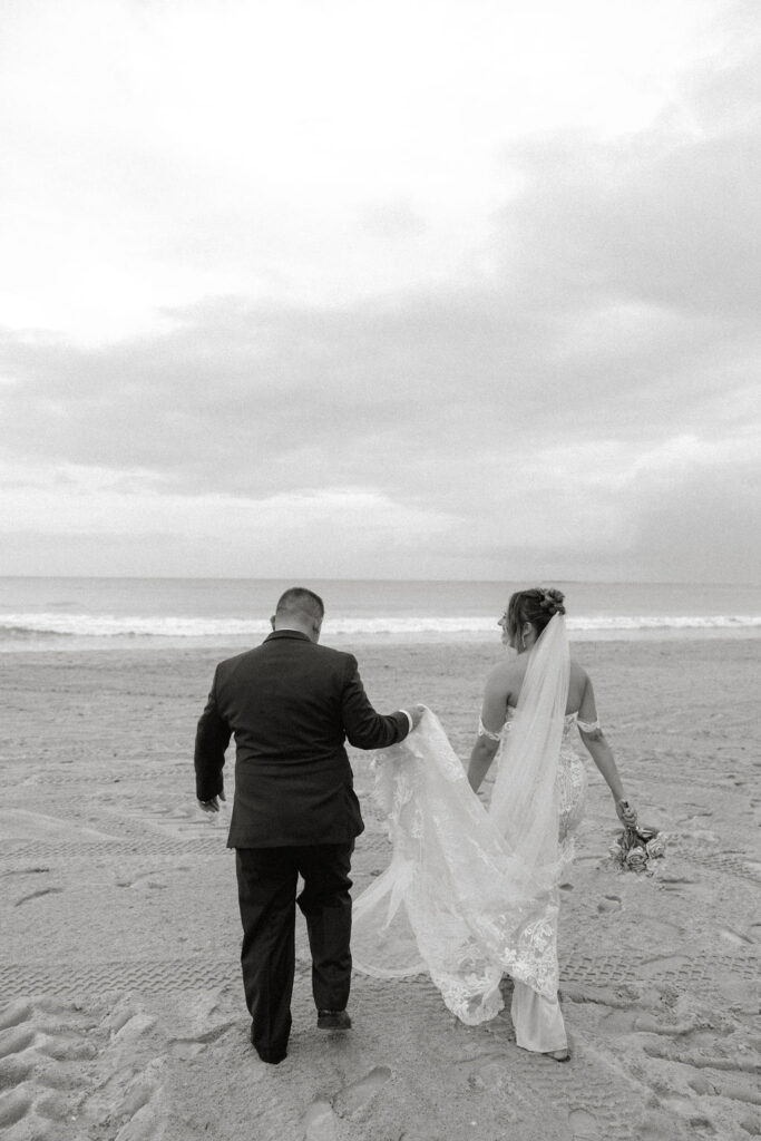 groom holding brides train of her wedding dress in the sand